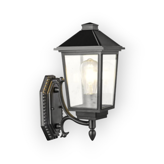 The image showcases a classic-style outdoor wall lantern with a sophisticated design. The lantern has a dark metal finish and features intricate detailing, including decorative ridges and finials. Clear glass panels are fitted into the lantern, allowing the light from within to shine brightly and illuminate the surroundings. This lantern is designed to add a touch of elegance to any exterior space, such as entryways, patios, or alongside pathways.