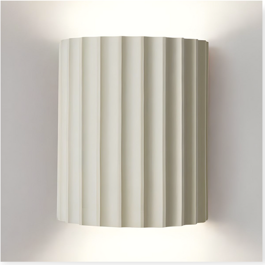 This image features the Eli Wall Light, which showcases a cylindrical form with elegant vertical ridges, creating a soft, diffused light that radiates from its surface. The pristine white finish adds a fresh, modern touch to any interior space.