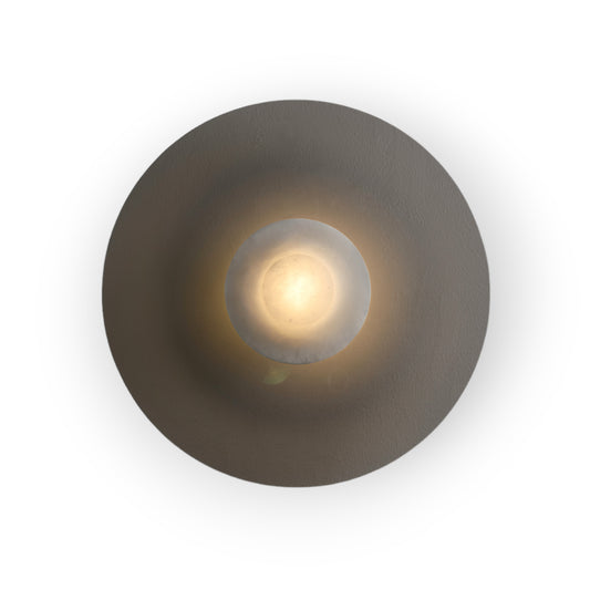 This image presents the Rhea Wall Light, with a central orb of luminous alabaster set against a backdrop of smoked glass, offering a soft and ambient glow. The interplay of materials creates a serene and luxurious lighting effect.